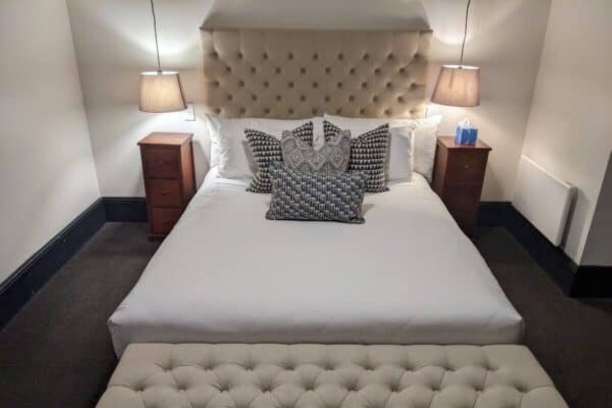 Un lit bien fait, doté d’une tête de lit capitonnée, deux tables de chevet avec des lampes et un pouf assorti.