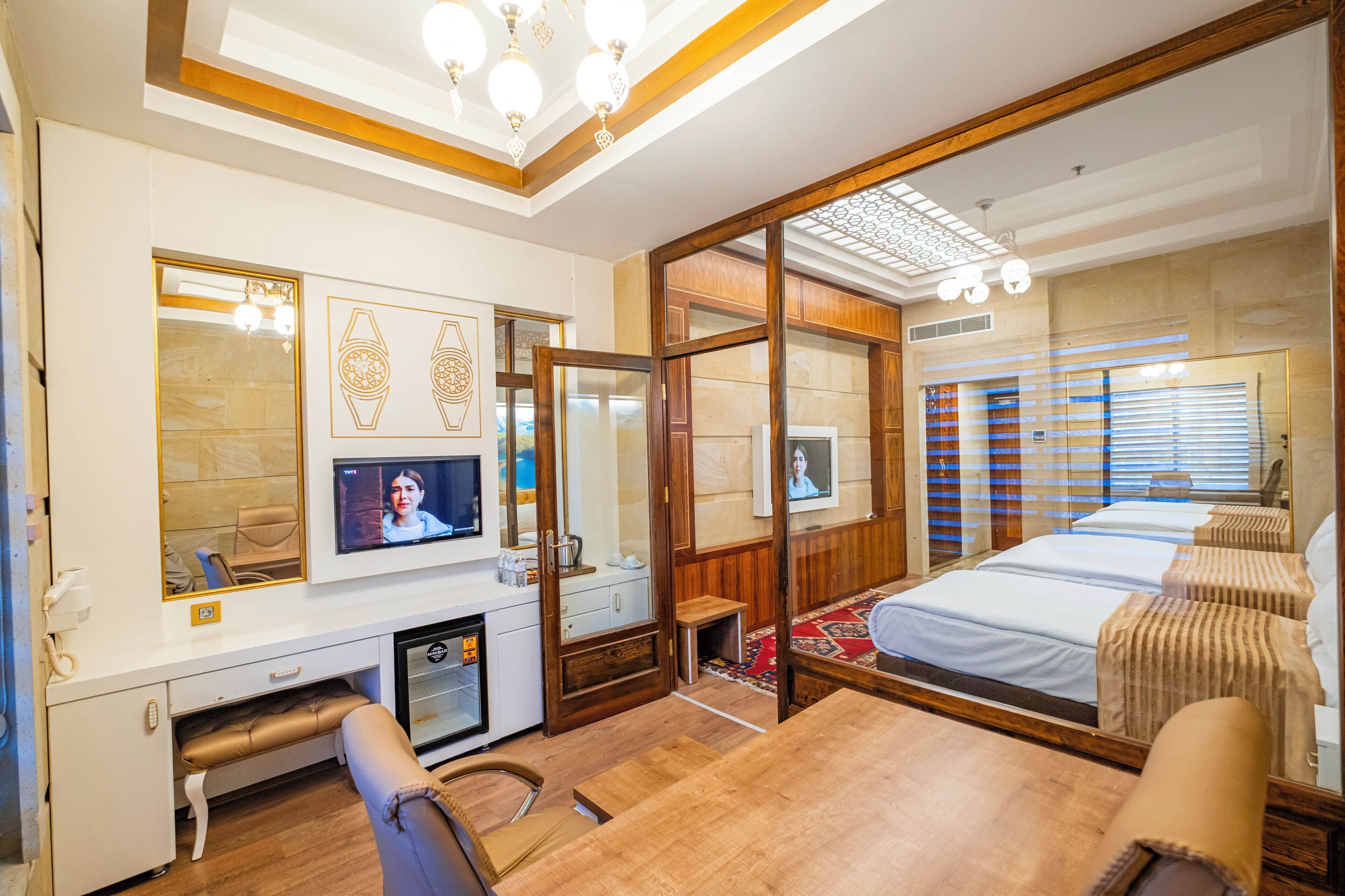 Habitació d'hotel amb sòl de fusta, una taula de treball amb un ordinador, un llit amb mantell de llit estampat i un mirall gran.