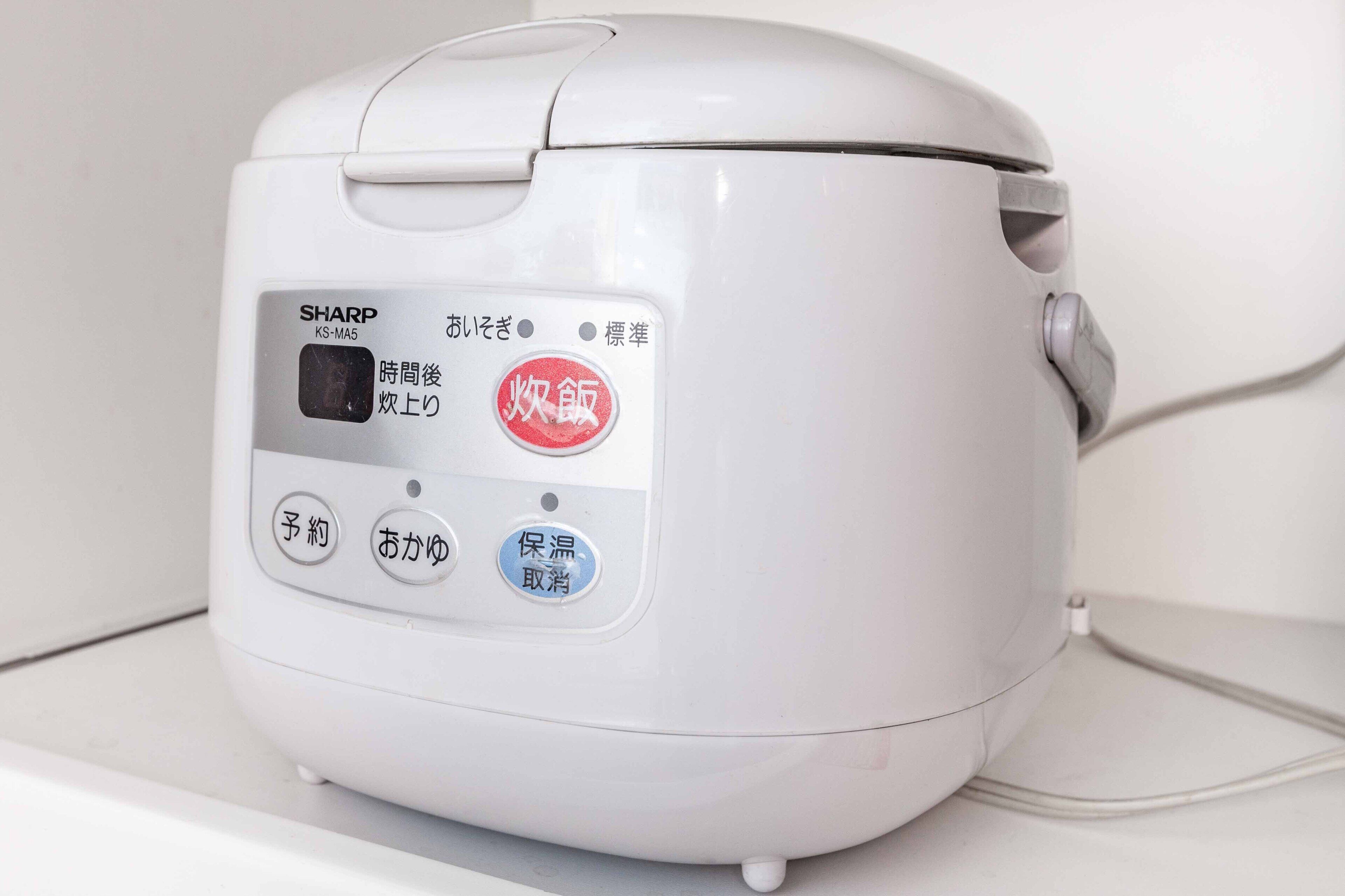 Een witte rijstkoker van SHARP met een digitaal display en meerdere knoppen, waaronder opties voor tijd, bereiding en instellingen.