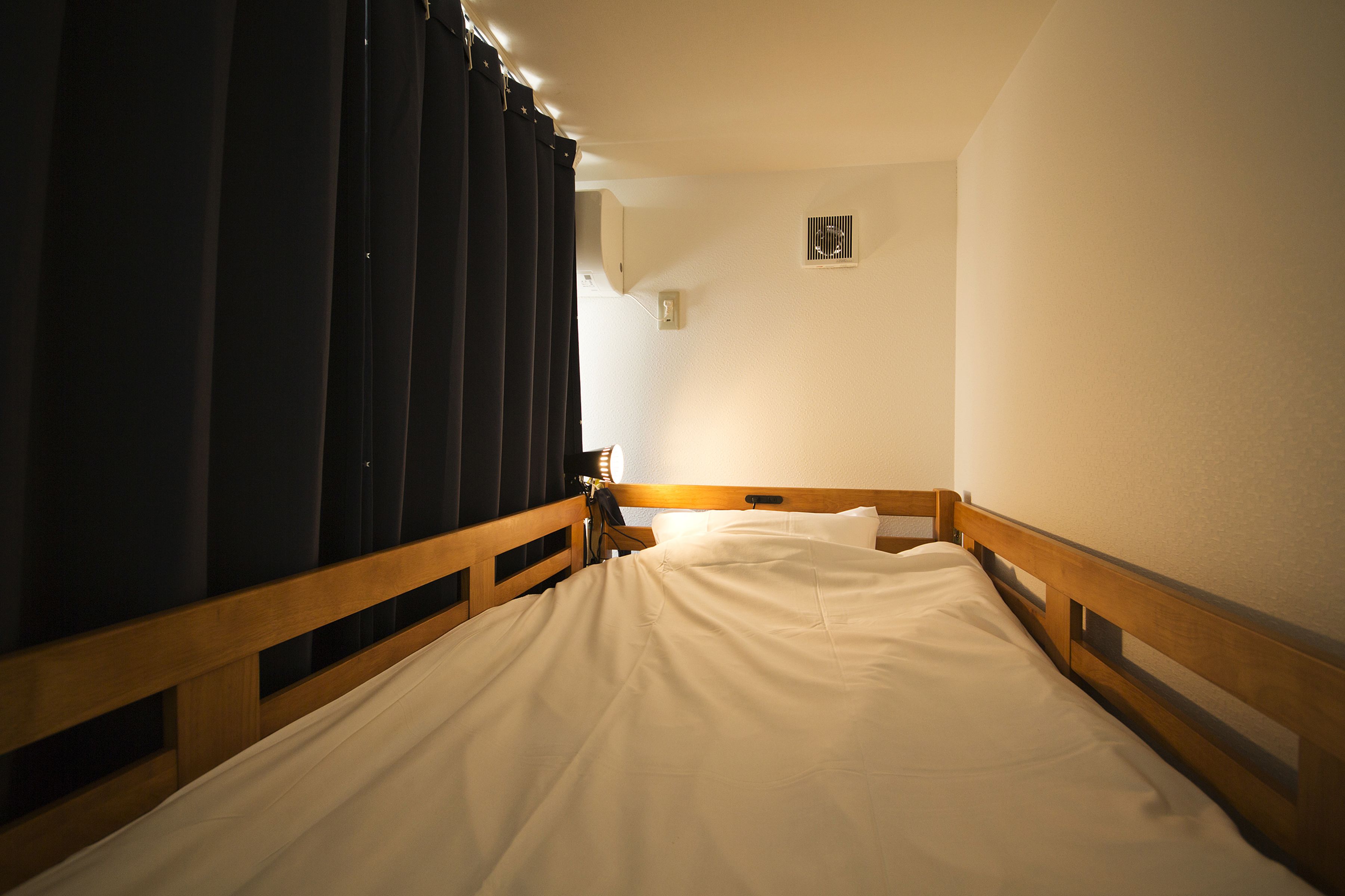 Una cama individual con cabecera de madera y ropa de cama blanca en una habitación con una cortina oscura en un lateral.