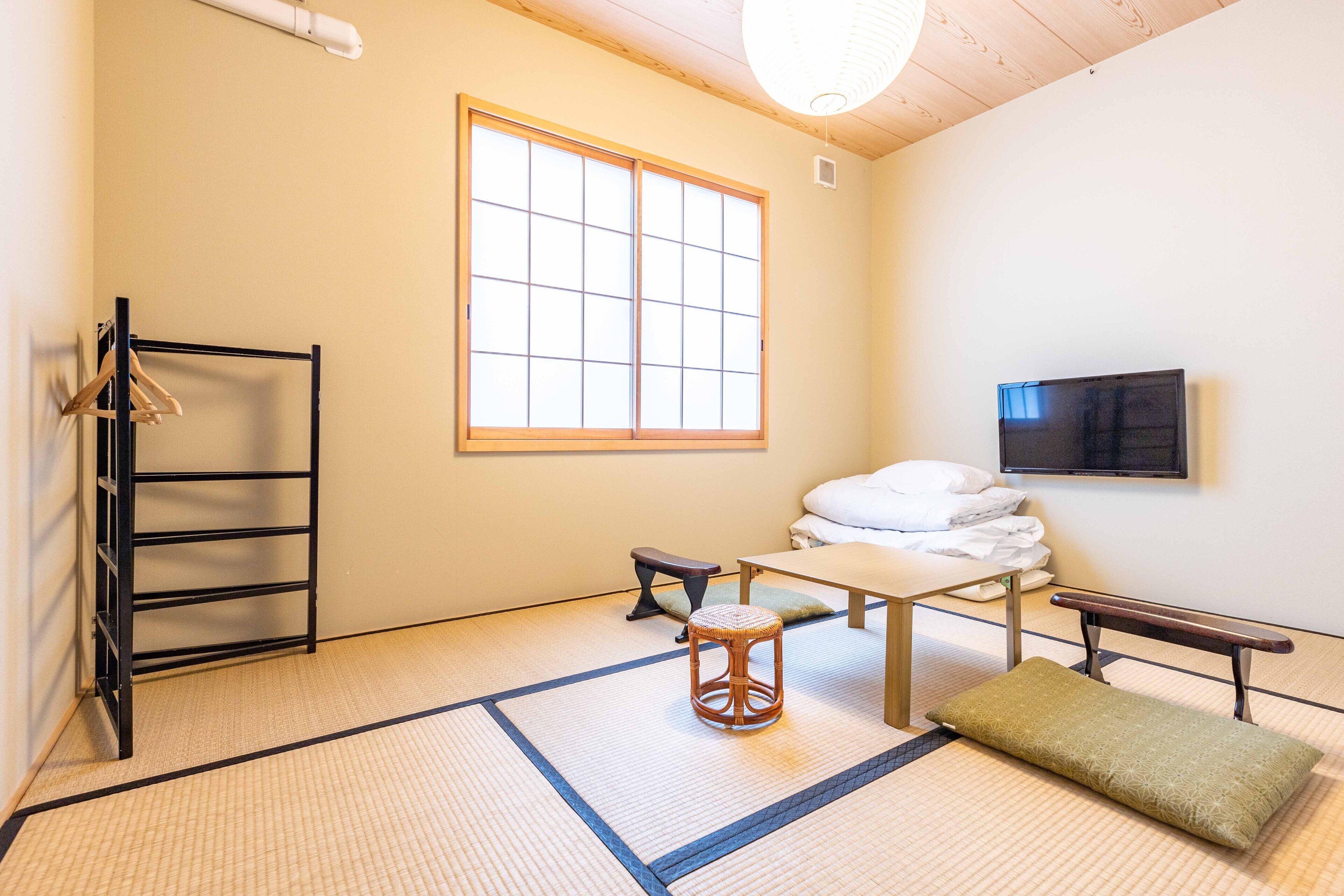 A room with tatami flooring, a low table with a cushion, a bench, a hanging clothes rack, and a large window.