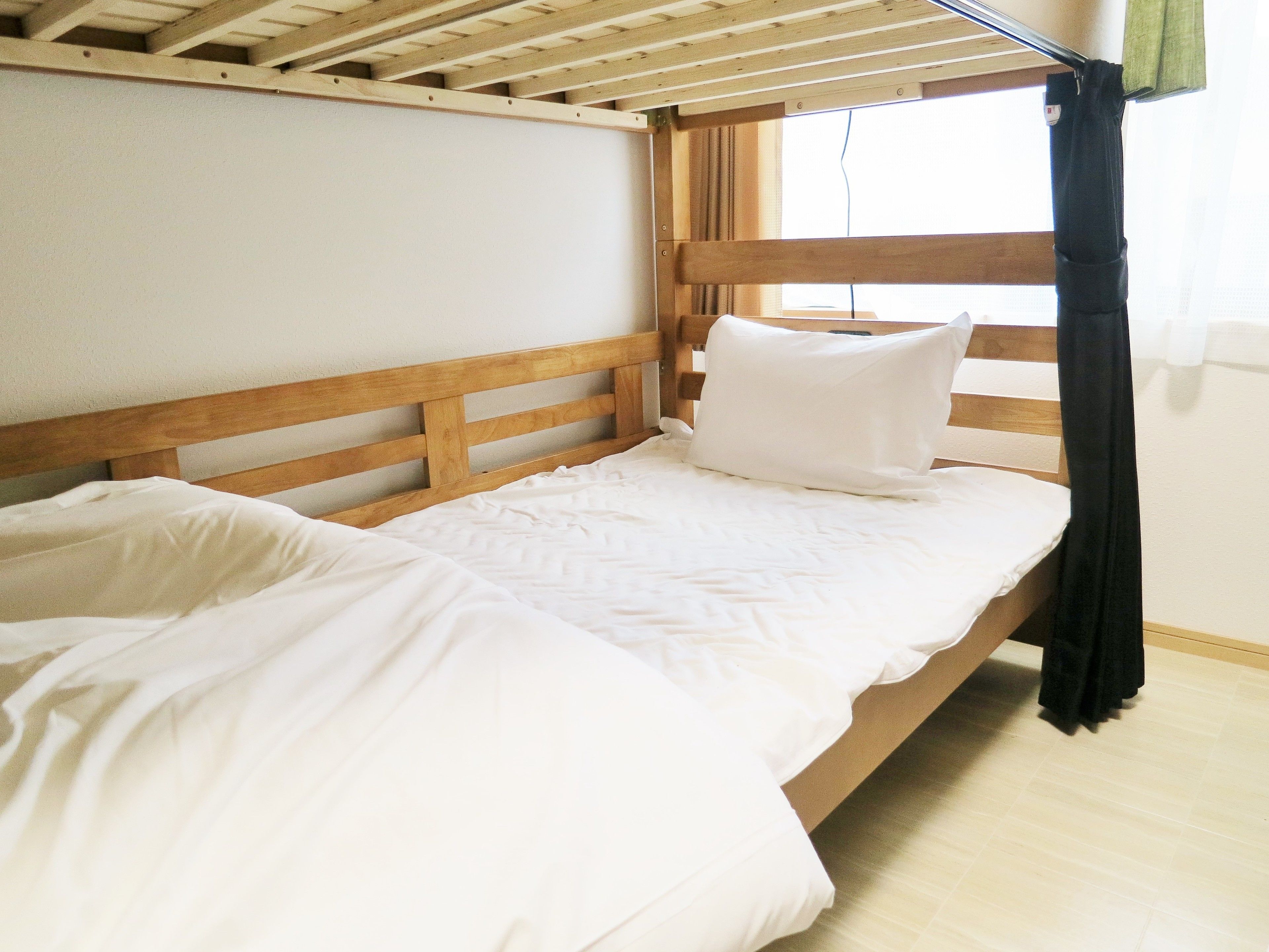 Een enkel bed met een houten hoofdeinde en wit beddengoed in een kamer met een houten vloer en een raam met gordijn.