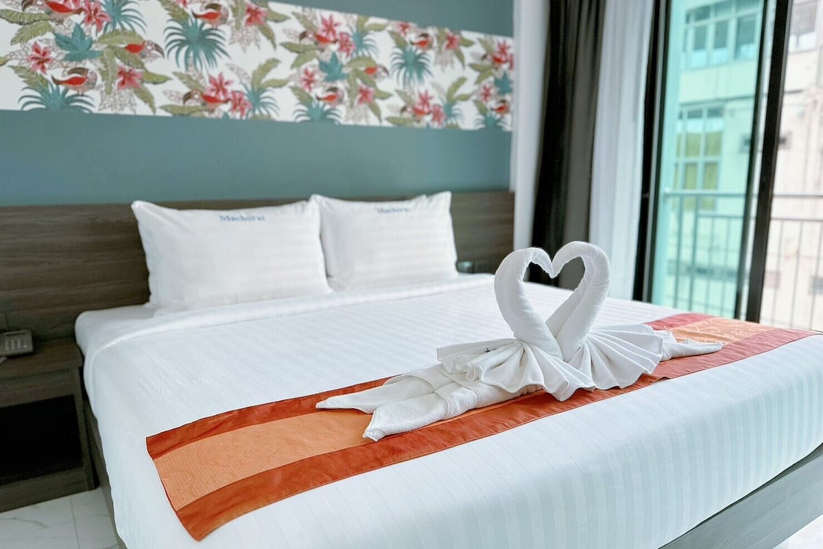 A hotel room with a neatly made bed, white bedding, and a heart-shaped towel decoration.