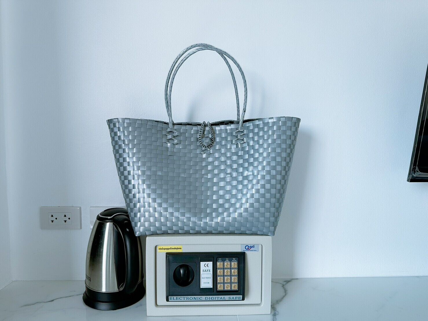 A silver woven bag on a scale next to a kettle and a digital safe.