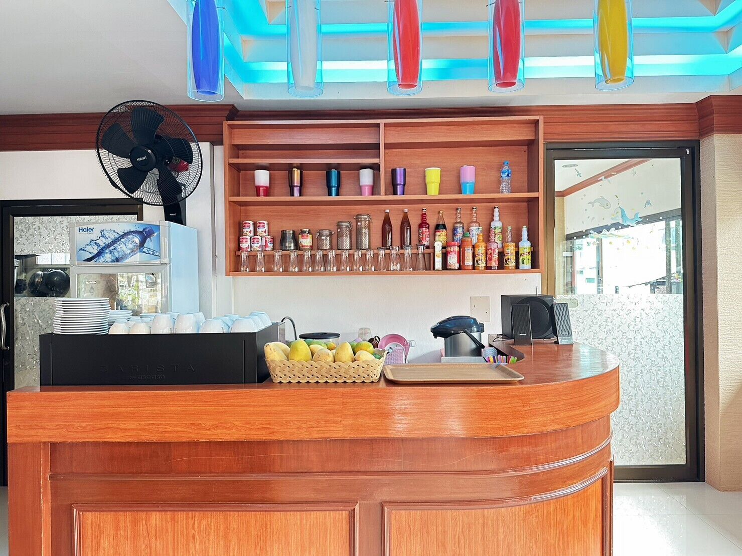 A hotel reception area with a wooden counter, a fan, a refrigerator, and a shelf with various bottles and containers.