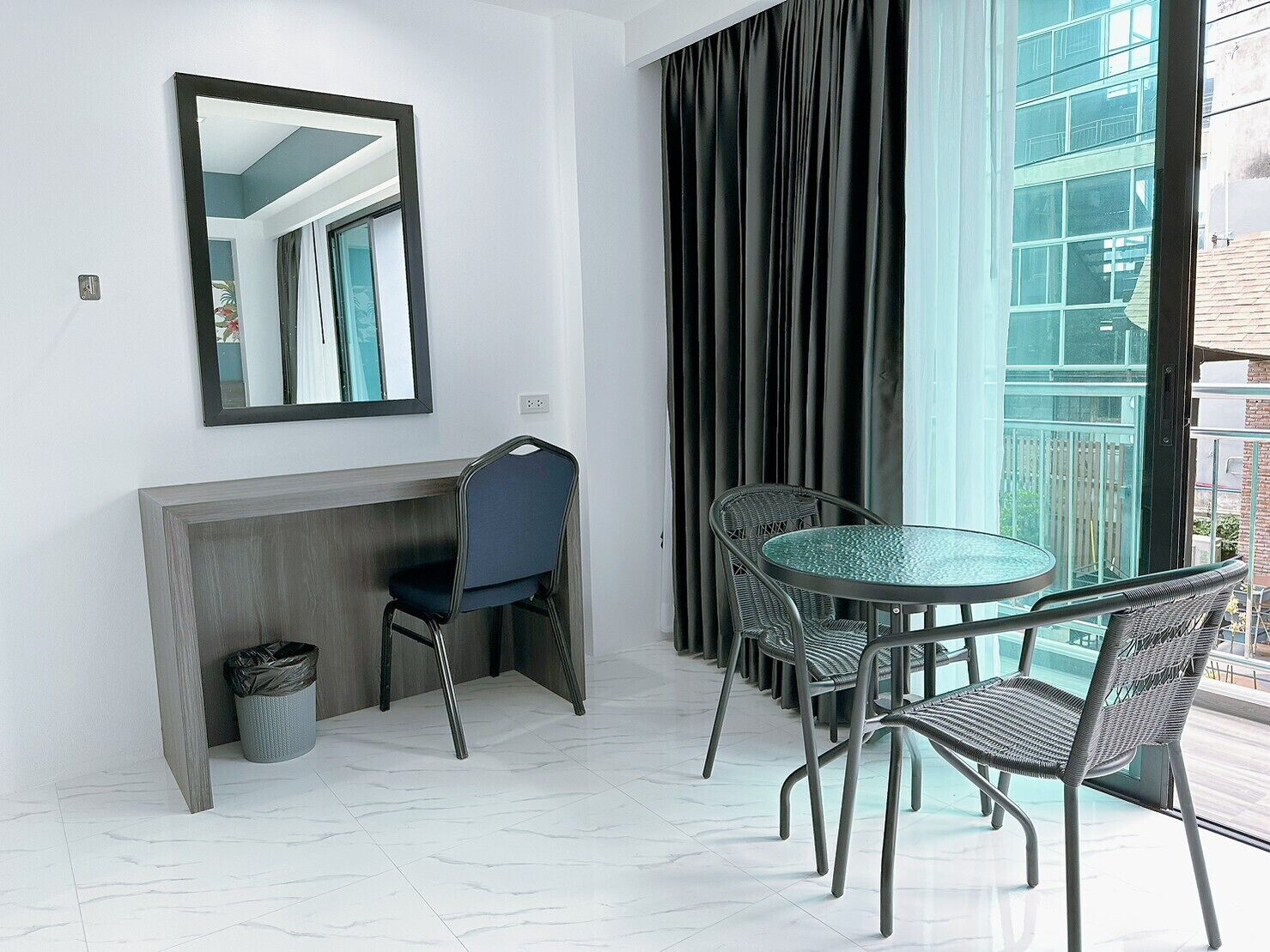 A modern room with a round glass table, four wicker chairs, a dark curtain, a large mirror, and a small trash bin.
