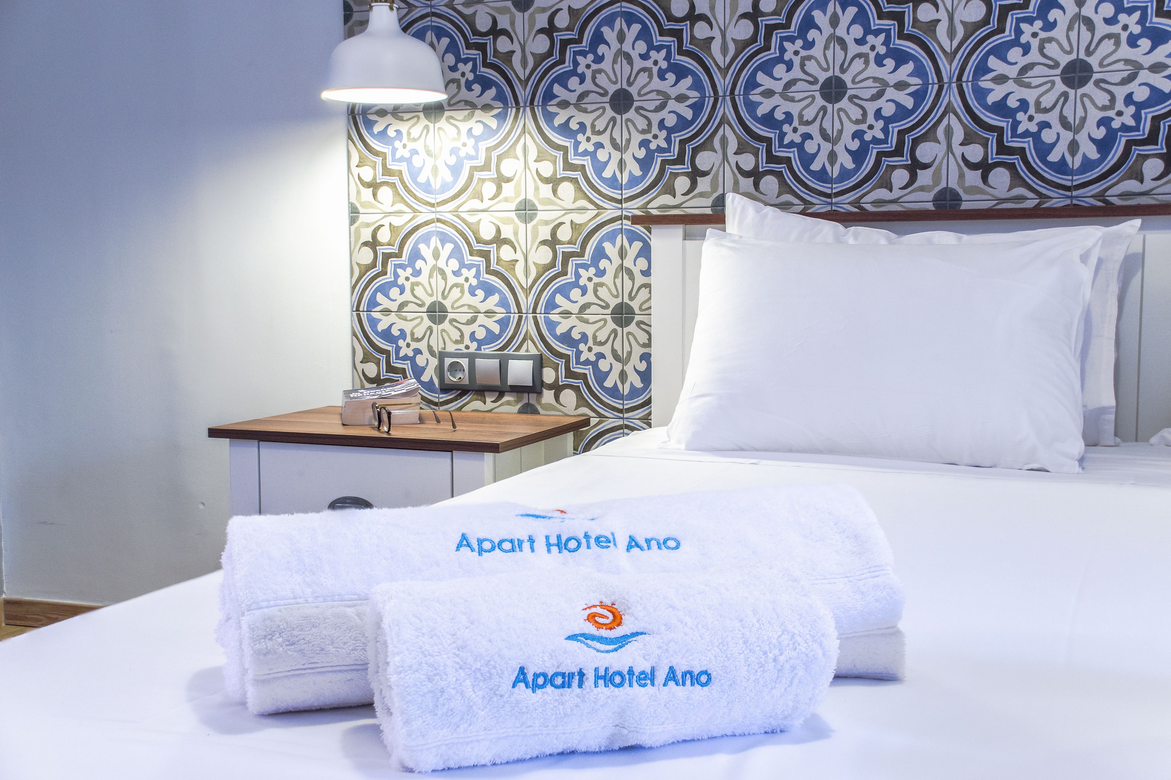 Une chambre d’hôtel avec un lit parfaitement fait, des serviettes blanches portant le nom de l’hôtel, une table de chevet avec une lampe et un mur décoré d’un motif.