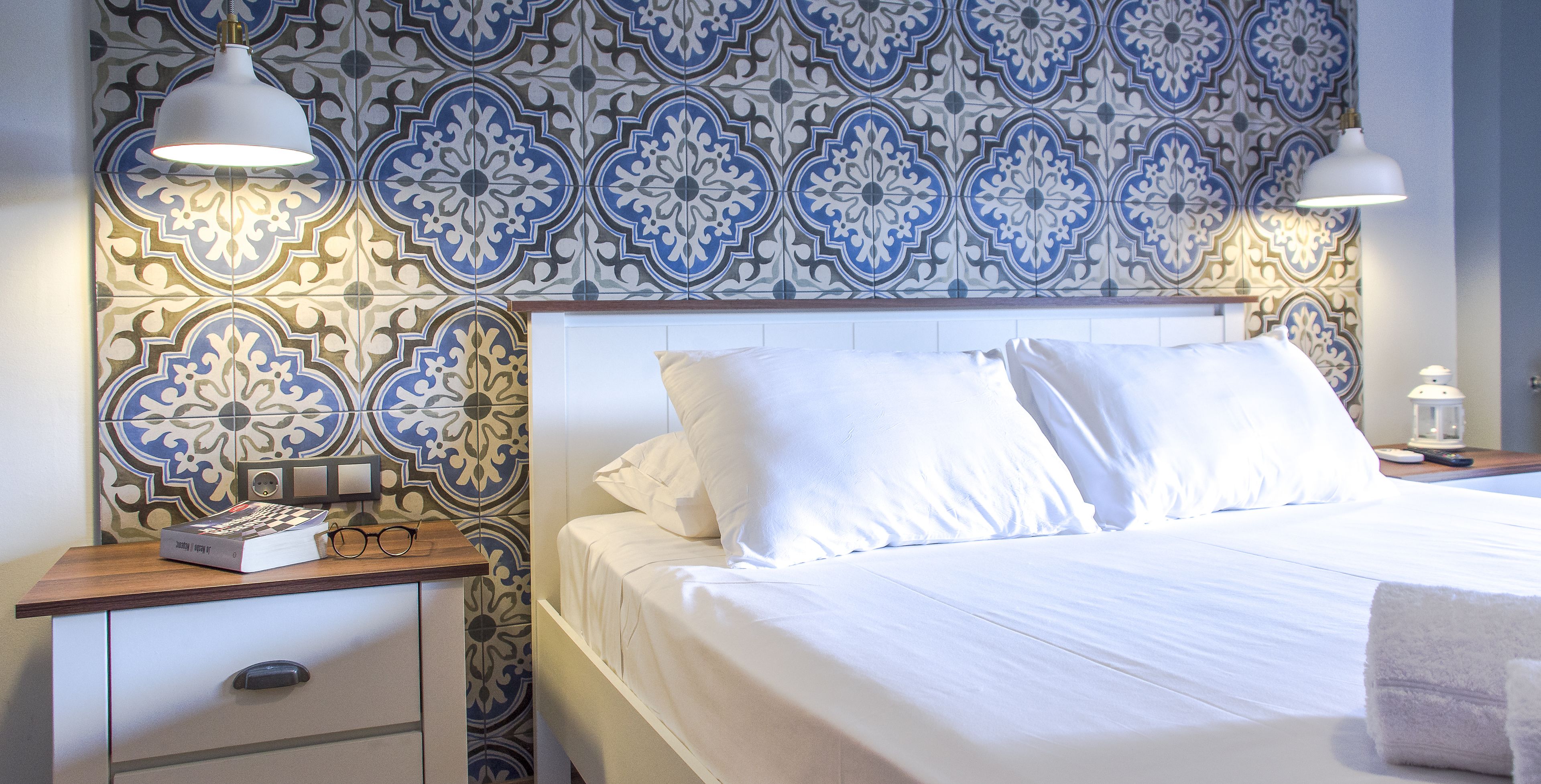 A neatly made bed with white linens, a bedside table with a book and glasses, and a patterned tile wall.