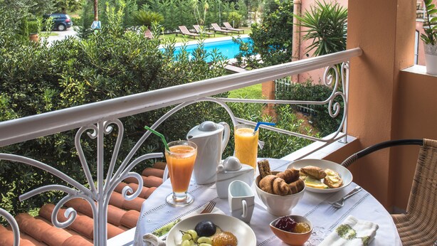 A balcony with a table set for breakfast, overlooking a pool and lush greenery. - Ano Aparthotel (Corfu)
