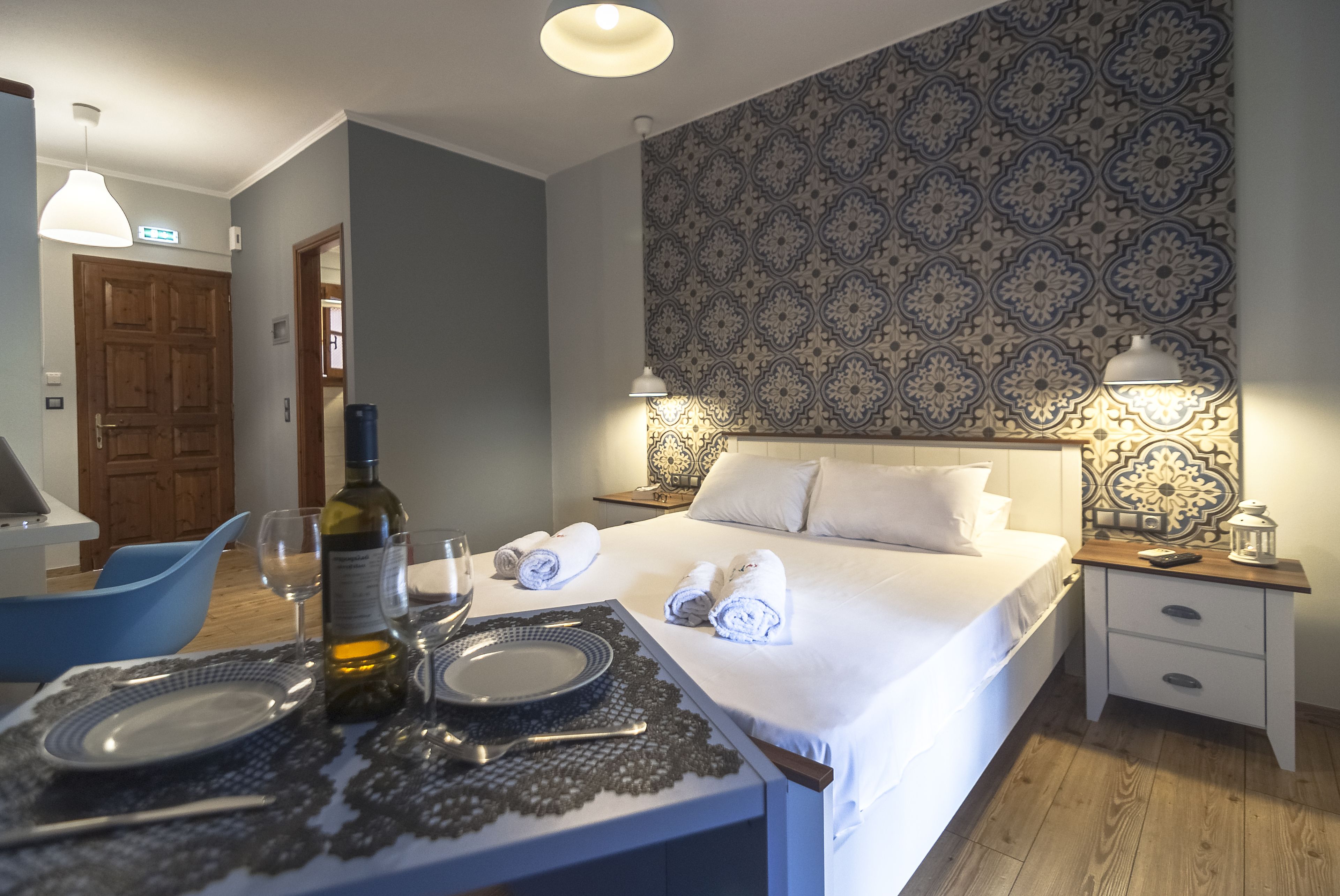 A hotel room with a neatly made bed, a bedside table with a lamp, a nightstand, and a dining area with a table set for two.
