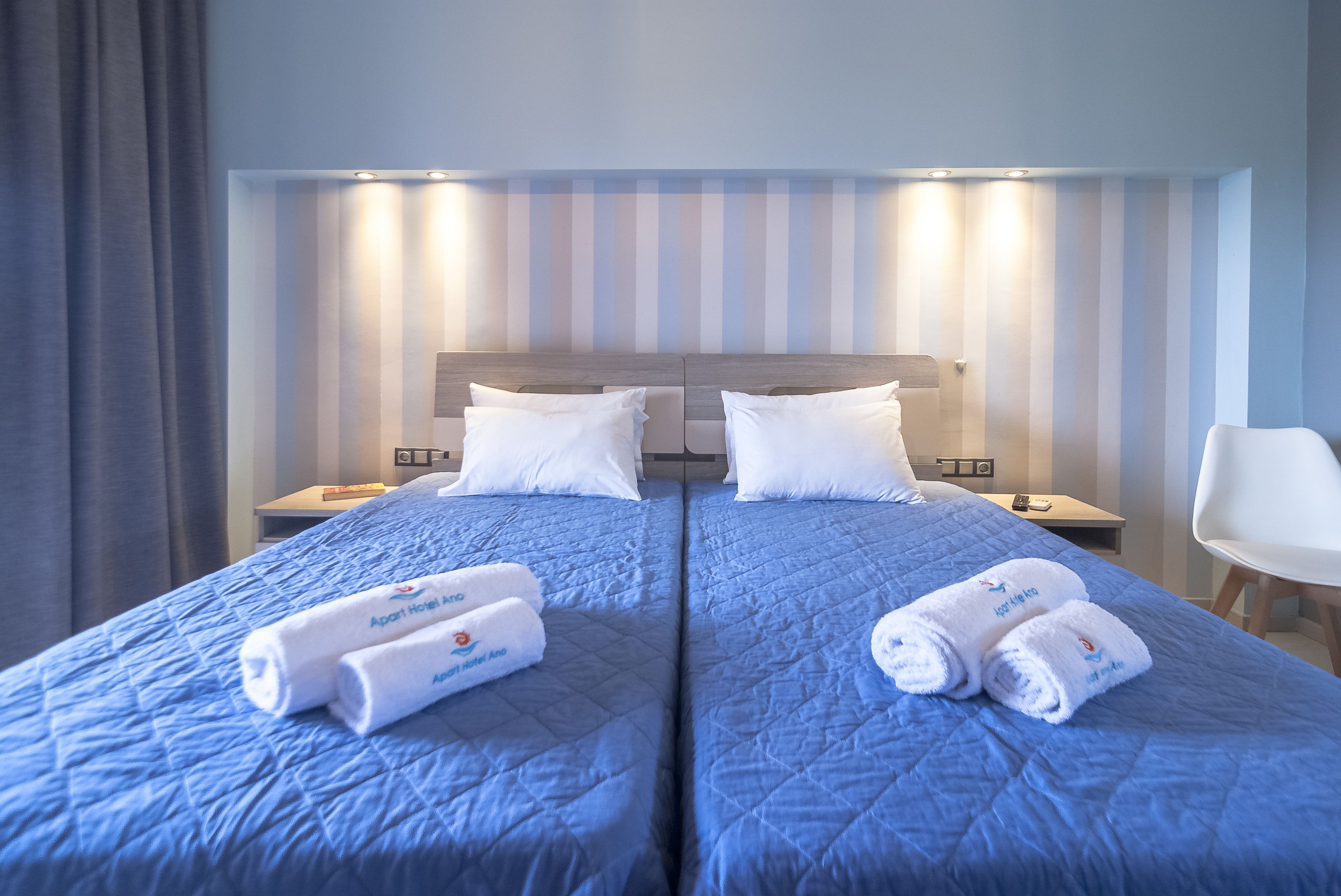 A hotel room with two beds, each with a blue quilted bedspread and white pillows. There are two sets of white towels with the hotel logo on them, and a white chair to the right.