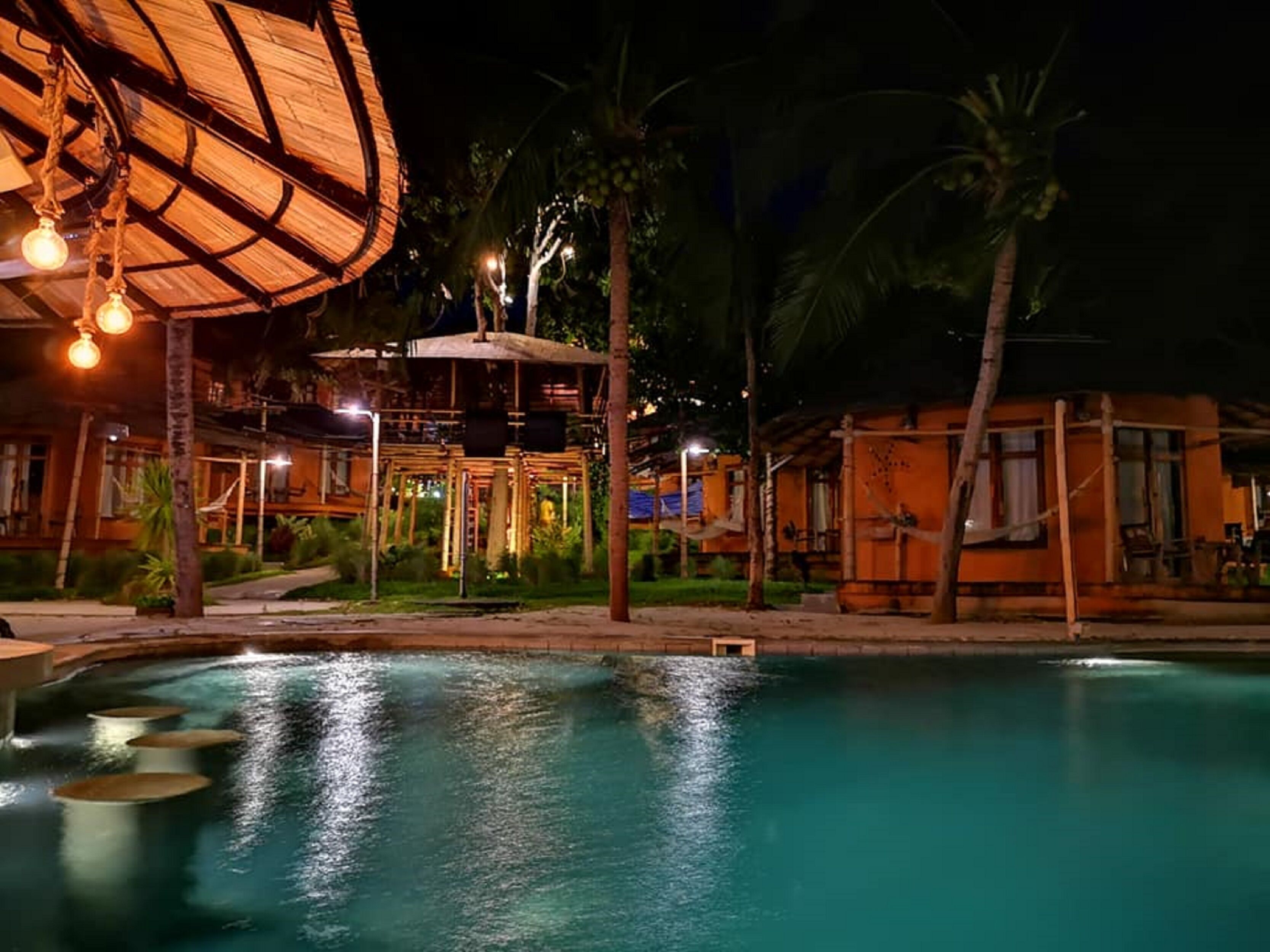 A nighttime view of a resort with a swimming pool, palm trees, and a wooden structure with hanging lights.