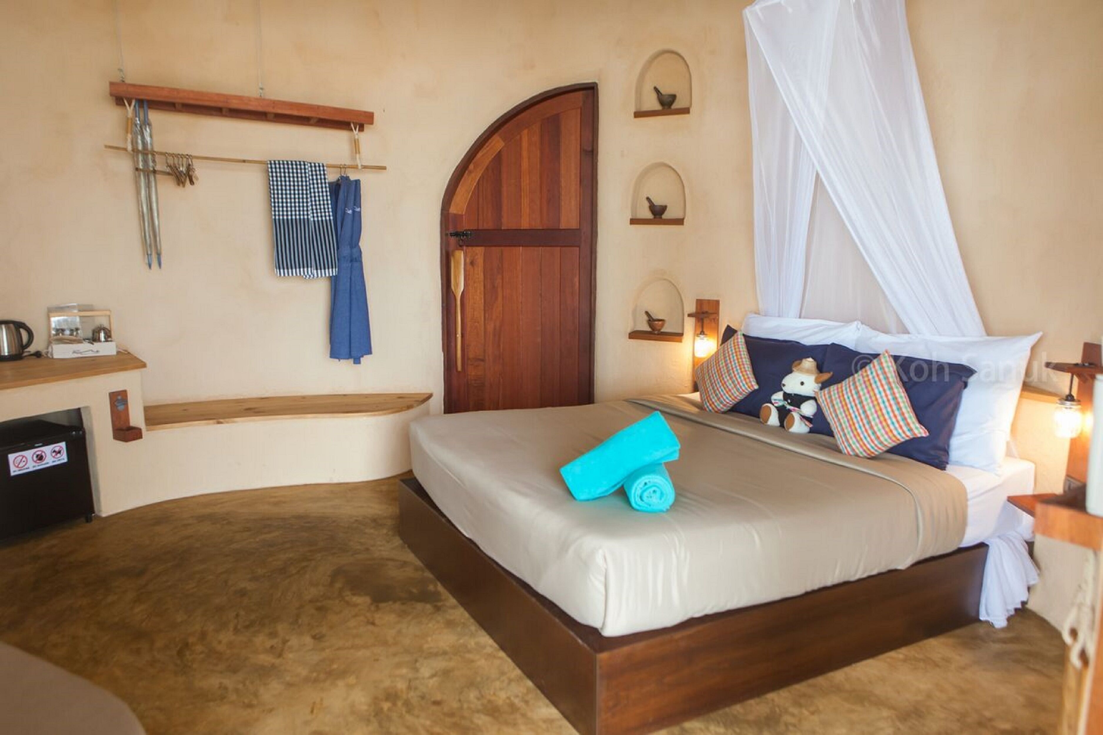 Une chambre à coucher avec un lit, une porte en bois, une étagère contenant des ustensiles de cuisine et une table de chevet avec une lampe.