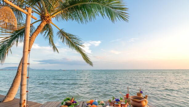 A wooden deck with a variety of dishes and traditional pottery, overlooking a calm sea with palm trees. - The Mud (Koh Samui)