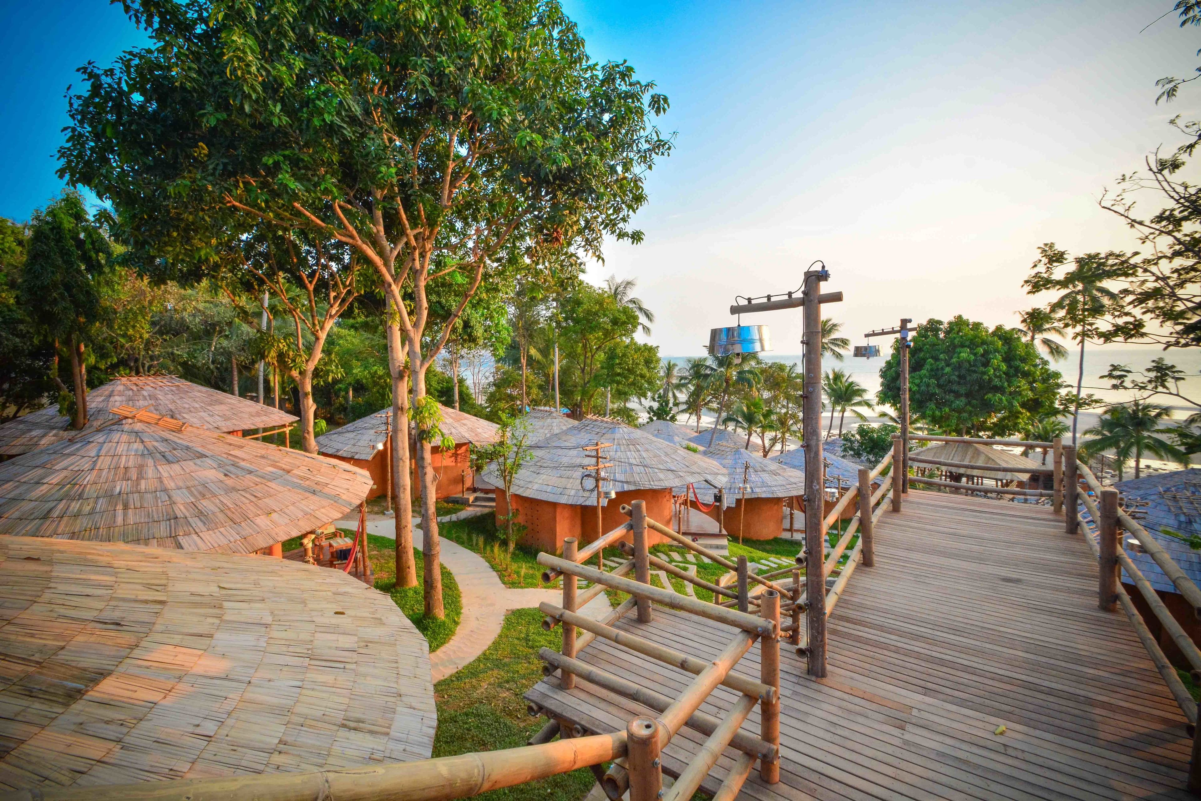A wooden deck leading to thatched-roof huts, surrounded by lush greenery and palm trees.