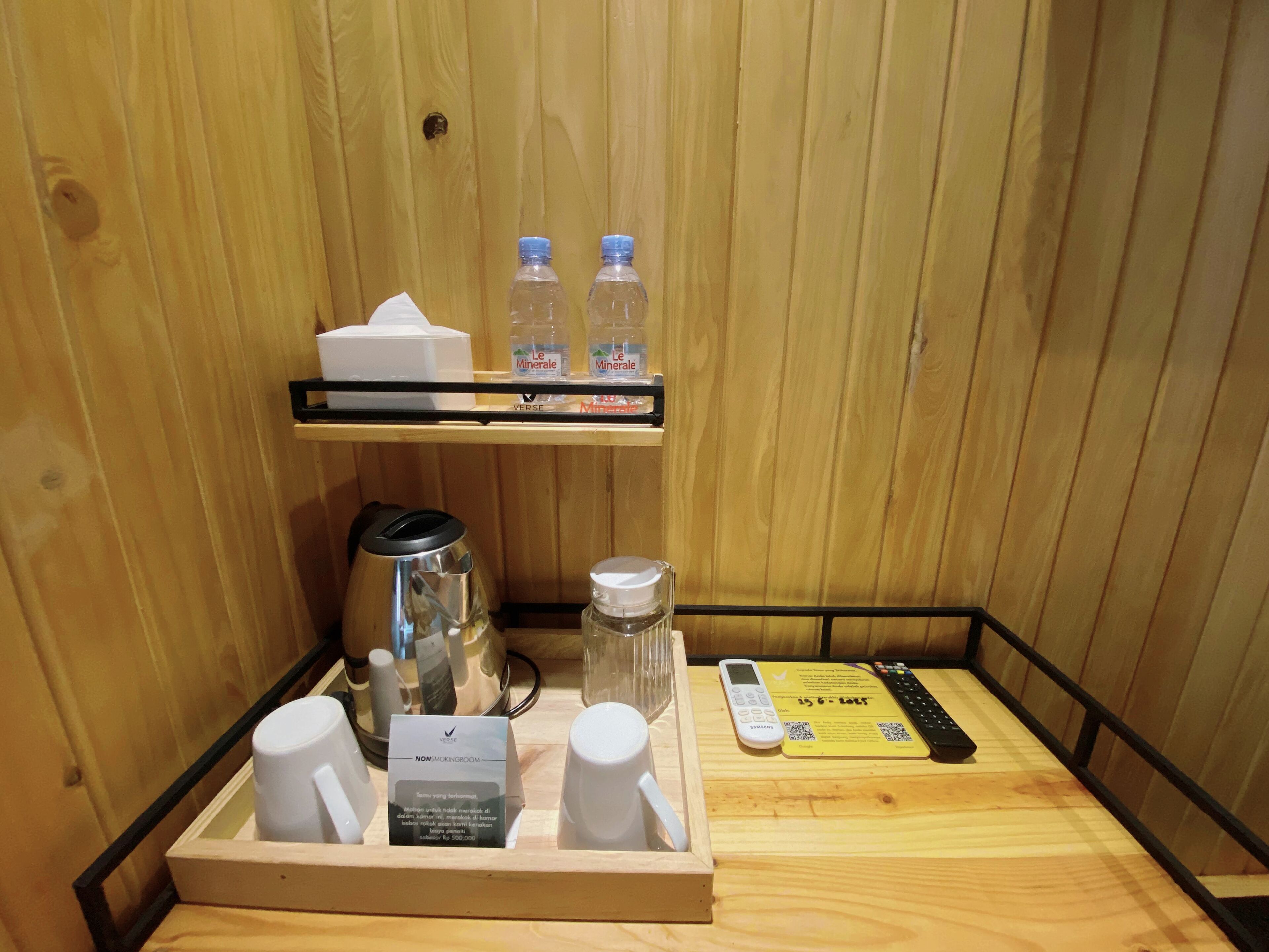 Una habitación de madera con una bandeja que contiene una cafetera, dos tazas blancas, una botella de agua y una caja de pañuelos. Hay un control remoto y una tarjeta sobre la mesa.