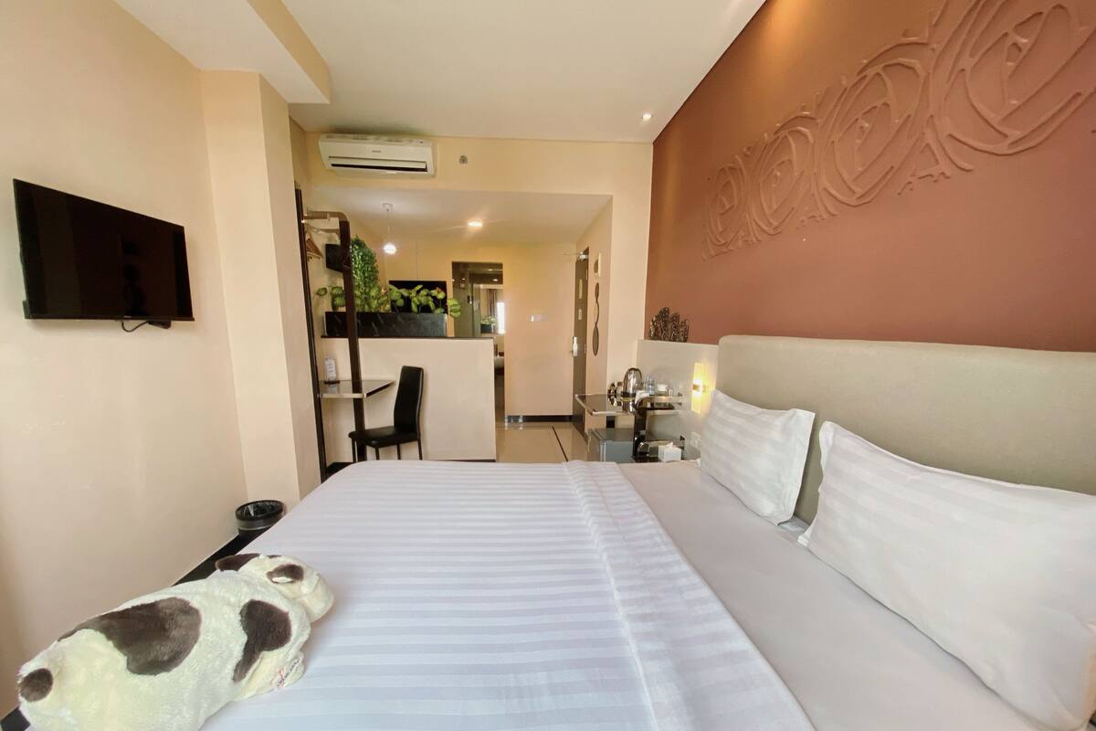 Habitación de hotel con una cama grande, un televisor de pantalla plana montado en la pared, un escritorio con una silla y una zona de kitchenette.