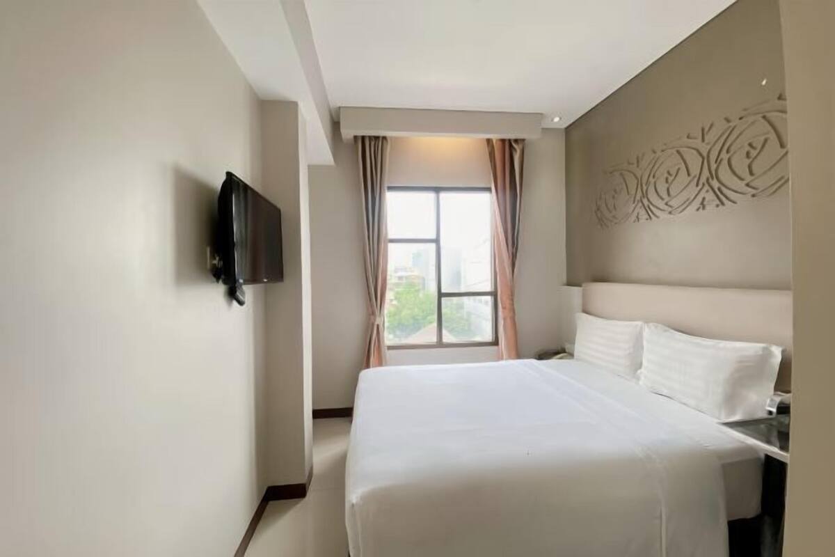 Habitación de hotel con cama, televisor montado en la pared, ventana con cortinas y un adorno de pared.