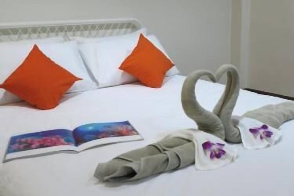 Una cama bien hecha con dos almohadas naranjas, un arreglo de toallas con forma de corazón, un libro y una almohada floral.