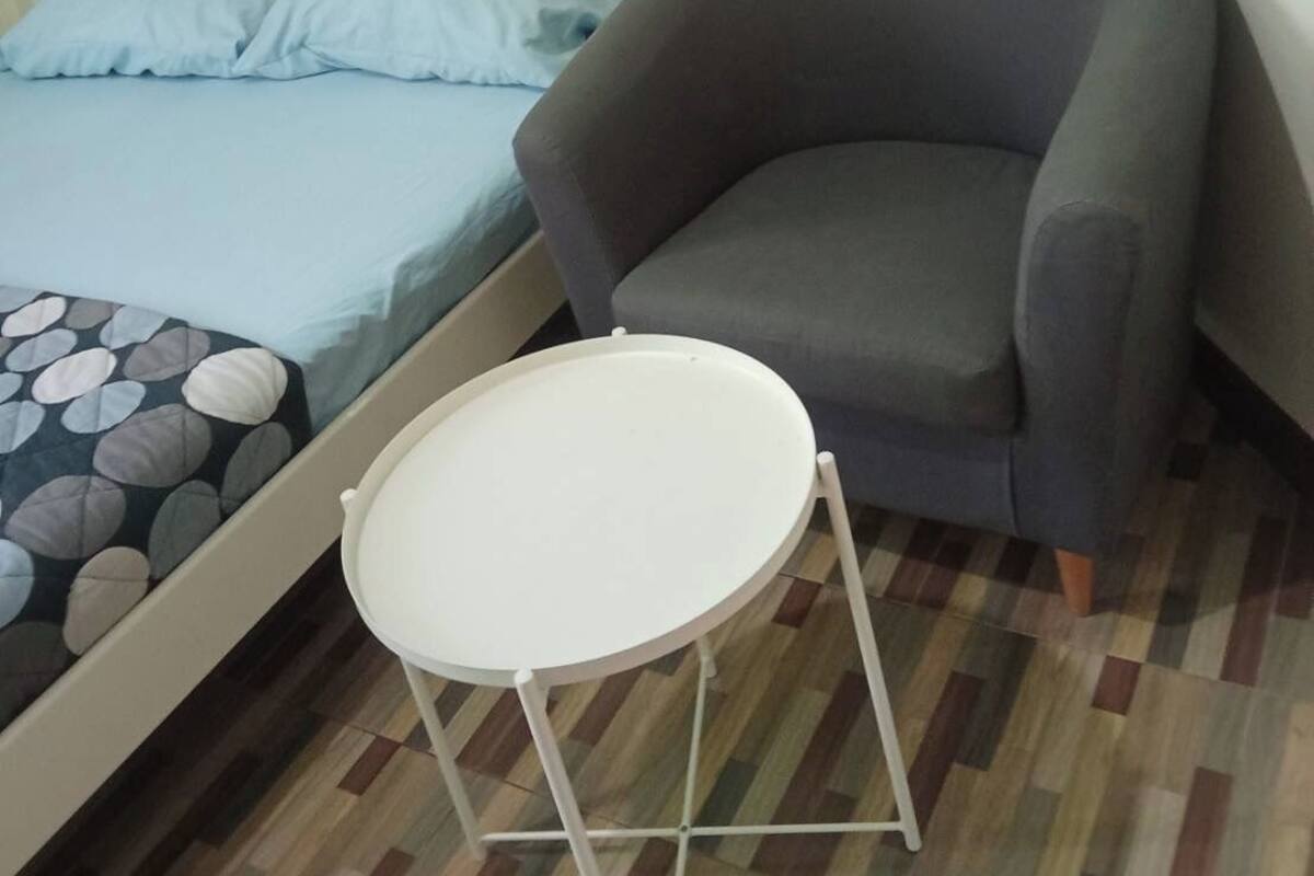Un dormitorio con una cama, un sillón gris y una pequeña mesa blanca.