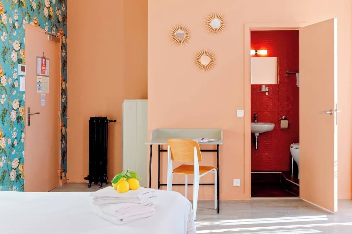 A hotel room with a bed, a desk, a chair, a bathroom with a red wall, and a door leading to another room.