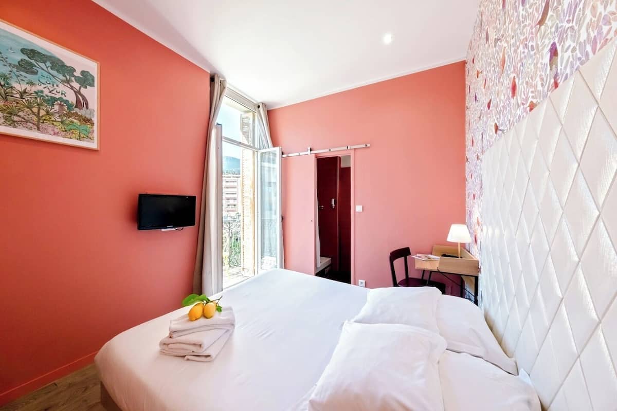 A hotel room with a bed, a desk with a chair, a television on the wall, and a window with curtains.