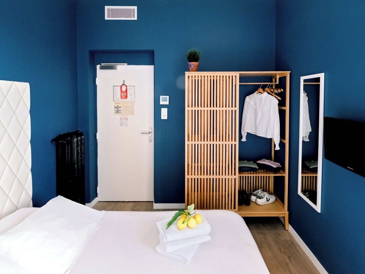 A hotel room with a bed, a wardrobe, a door with a fire alarm, and a small plant on top of the wardrobe.