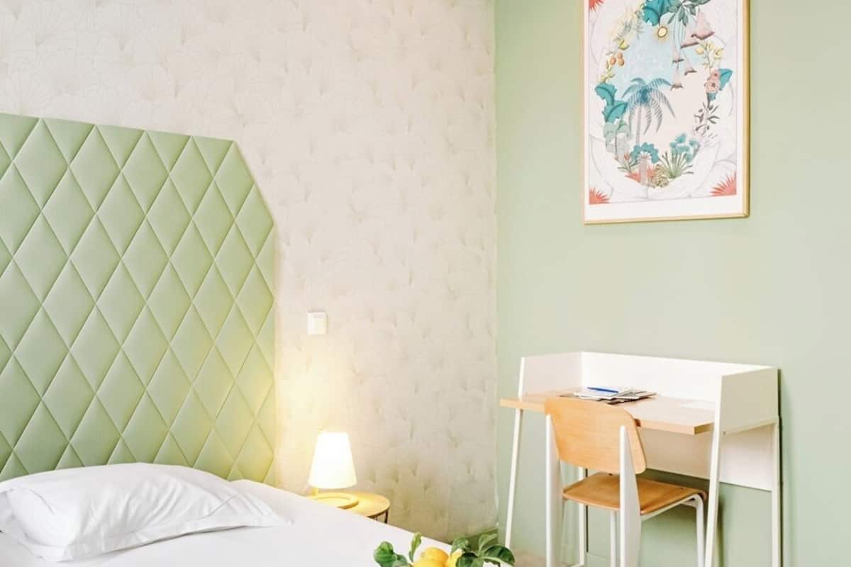 A neatly made bed with a green headboard, a desk with a chair, and a framed floral artwork on the wall.