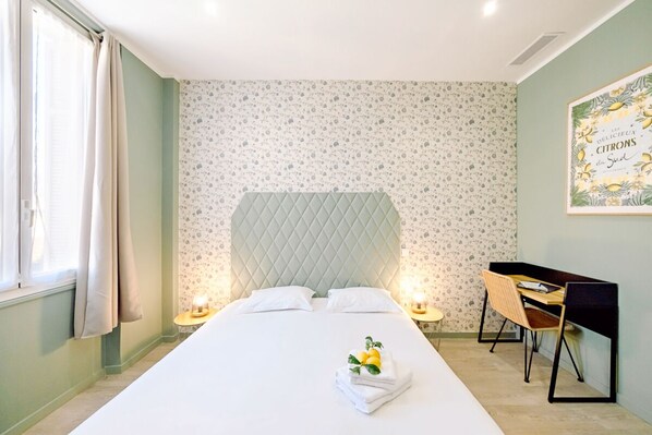 A hotel room with a bed, a desk, and a chair. There is a window with curtains, a wall with a floral pattern, and a framed picture on the wall.