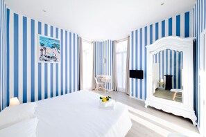 A hotel room with striped blue and white walls, a bed with white linens, a bedside table with a lamp, a small table with a chair, and a large mirror with a white frame.