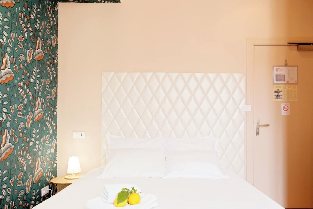 A hotel room with a quilted headboard, a bed with white linens, a nightstand with a lamp, and a door with a sign.