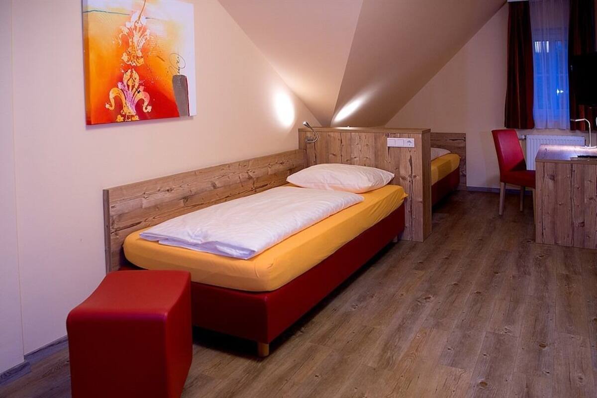 Quarto com piso de madeira, cama com estrutura vermelha, um banquinho vermelho, uma escrivaninha com uma cadeira e um quadro na parede.