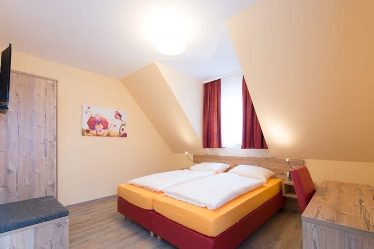 Um quarto com uma cama, um banco de madeira, um quadro e uma janela com cortinas.