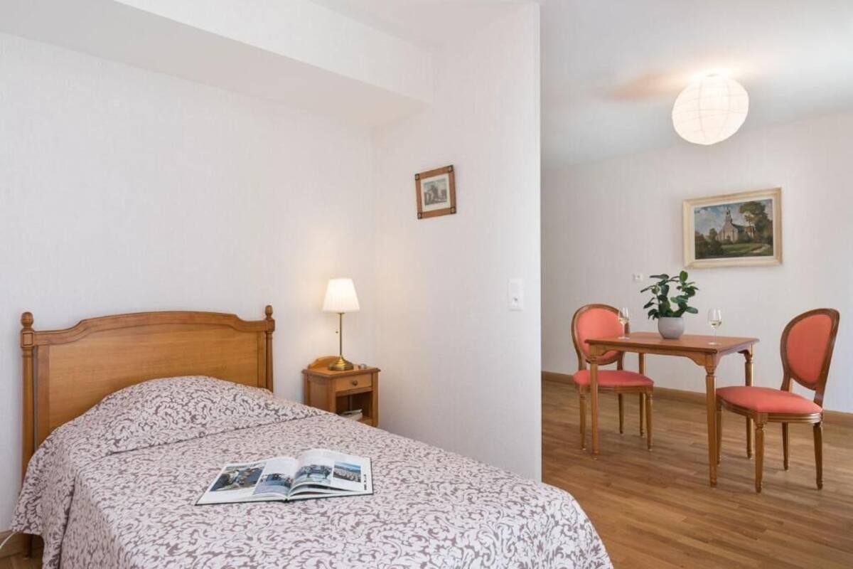 A bedroom with a wooden bed, a bedside table with a lamp, a small table with two chairs, and a potted plant.