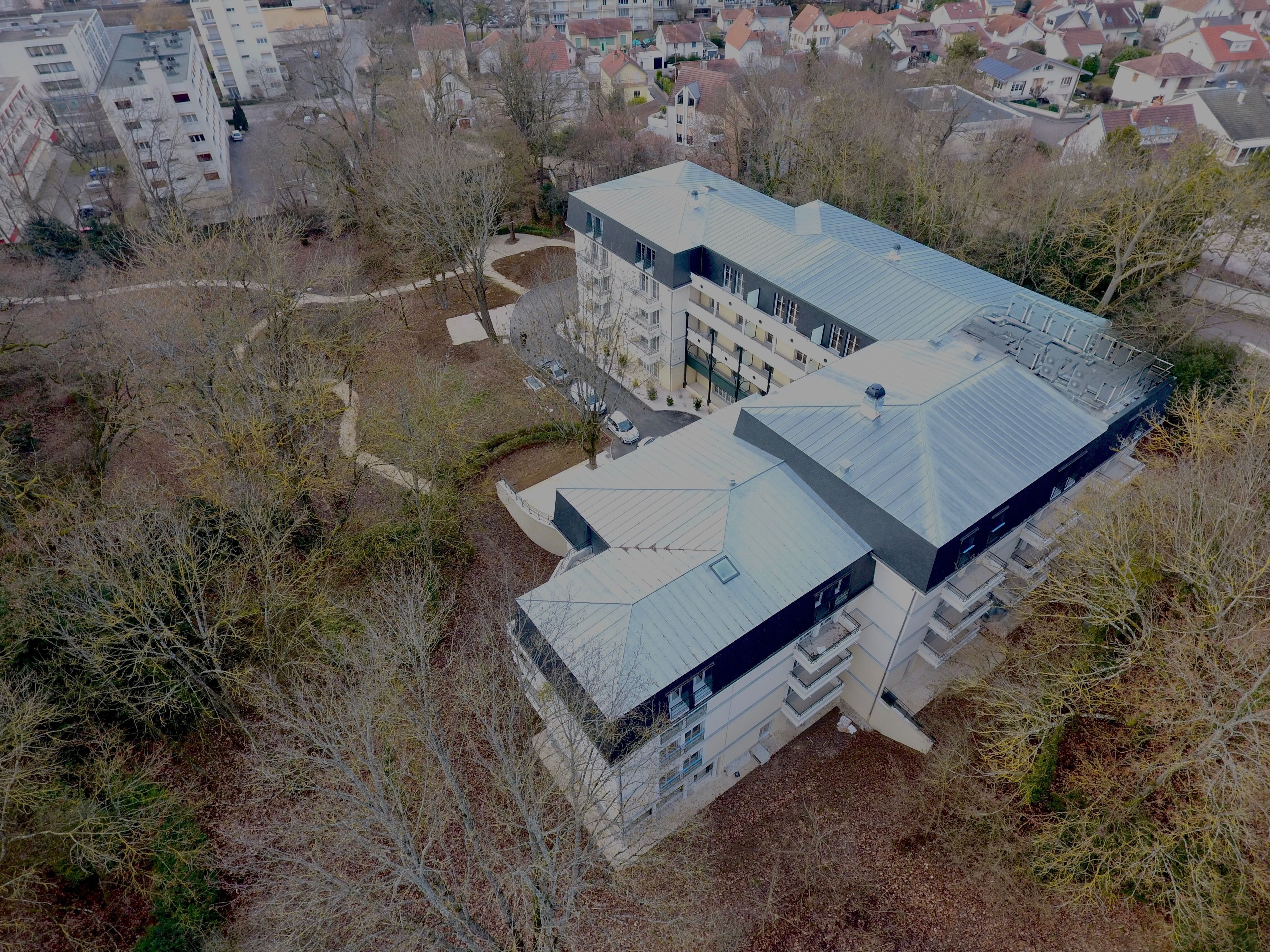 Vue aérienne d’un immeuble moderne à plusieurs étages, doté d’un toit plat, entouré d’arbres et d’autres immeubles résidentiels.
