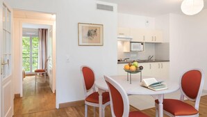 A dining area with a table and four chairs, a fruit bowl on the table, and a kitchen in the background. - Home Médicis Les Petites Roches (Dijon)