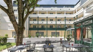 An outdoor cafe with tables and chairs, a large tree, and a building with balconies in the background. - Home Médicis Les Petites Roches (Dijon)