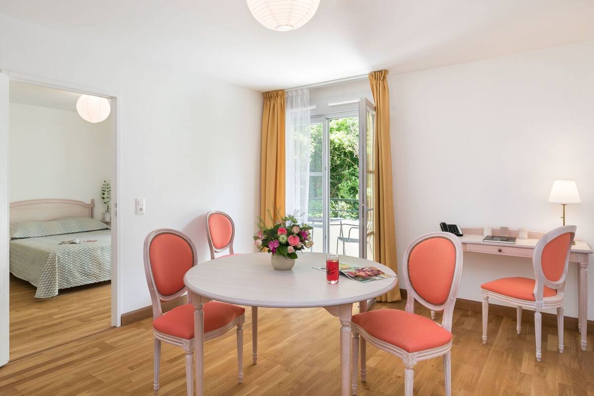 A room with a round table, four chairs, a desk, and a bed.