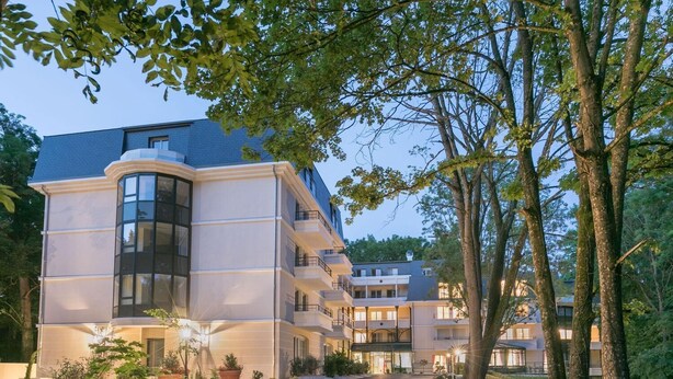 A multi-story hotel with a curved glass facade, surrounded by lush greenery and trees. - Home Médicis Les Petites Roches (Dijon)