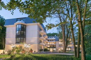 A multi-story hotel with a curved glass facade, surrounded by lush greenery and trees. - Home Médicis Les Petites Roches (Dijon)