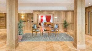 A spacious lobby with wooden flooring, two armchairs, a coffee table, and a dining area with a red curtain backdrop. - Home Médicis Les Petites Roches (Dijon)