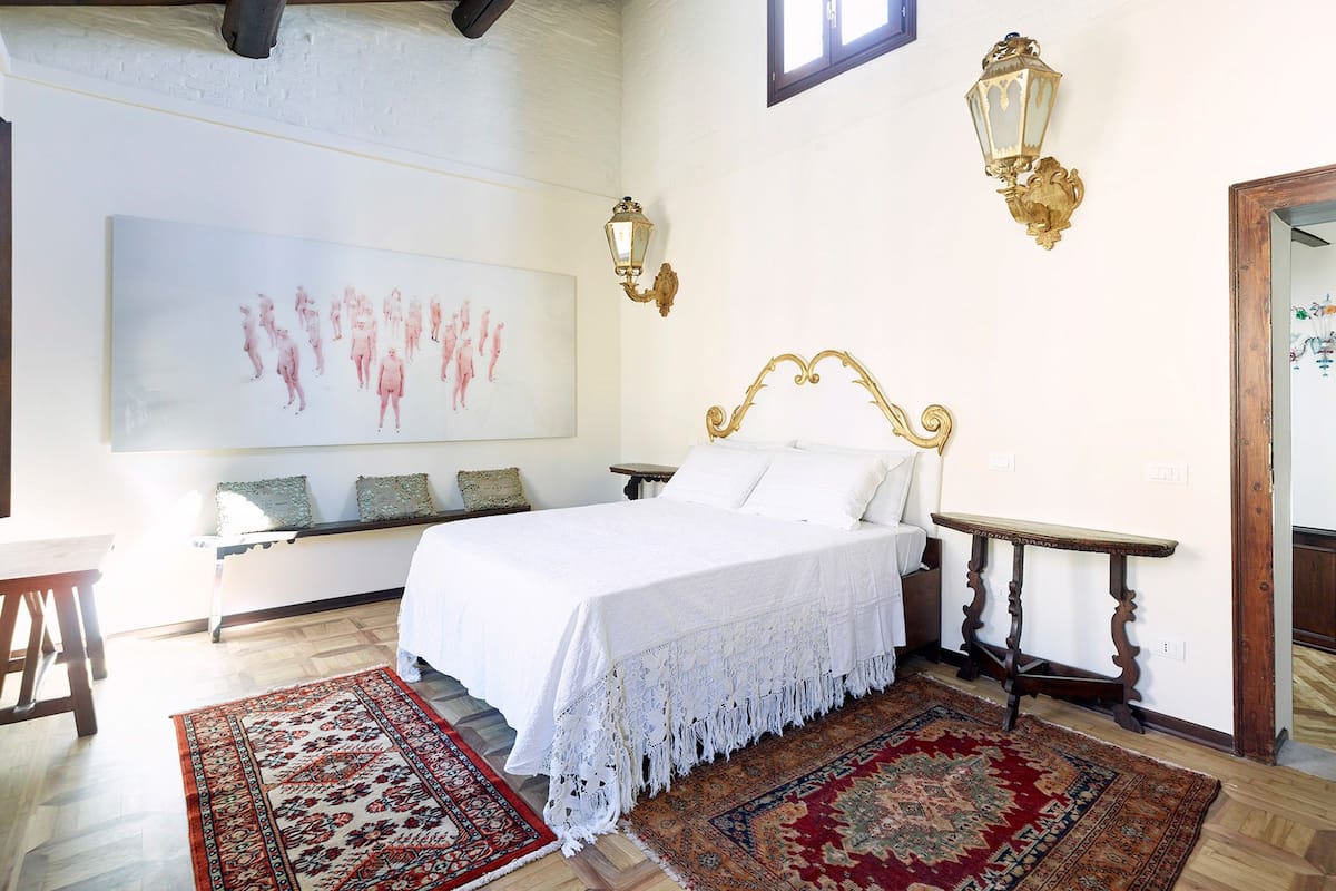 Une chambre à coucher avec un grand lit, deux tables de chevet, un banc avec des coussins, un grand tableau et deux lampes fixées au mur.