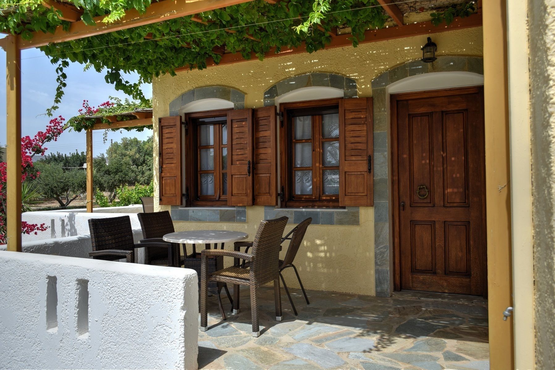 A covered outdoor seating area with wicker chairs and a table, stone flooring, and a pergola with climbing plants.