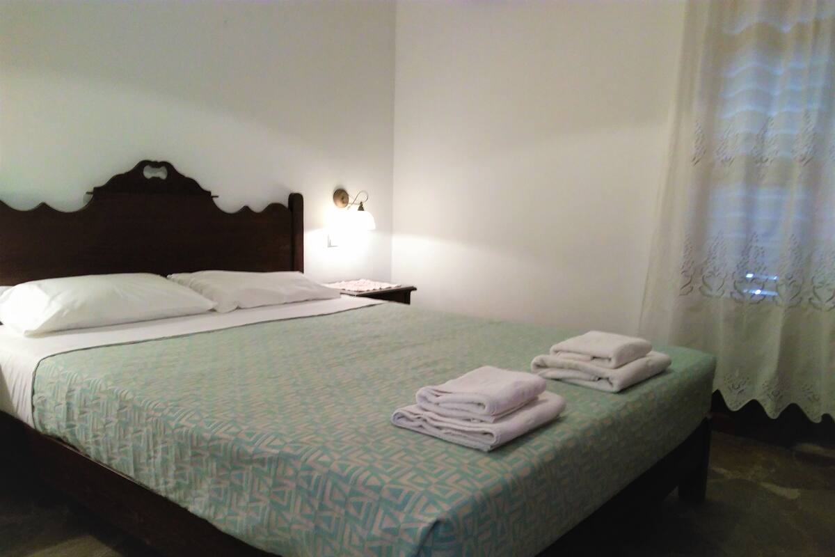 A neatly made bed with a patterned bedspread, pillows, and folded towels on a wooden bed frame in a room with white walls and a nightstand with a lamp.