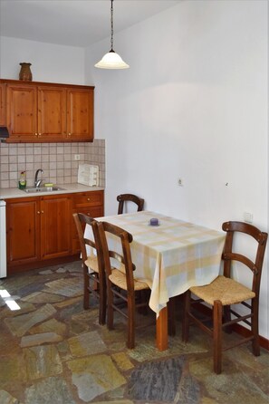 A dining area with a checkered tablecloth, wooden chairs, a hanging light fixture, and a kitchen with wooden cabinets and a tiled backsplash. - Grandes Apartments Kouremenos (Sitia)