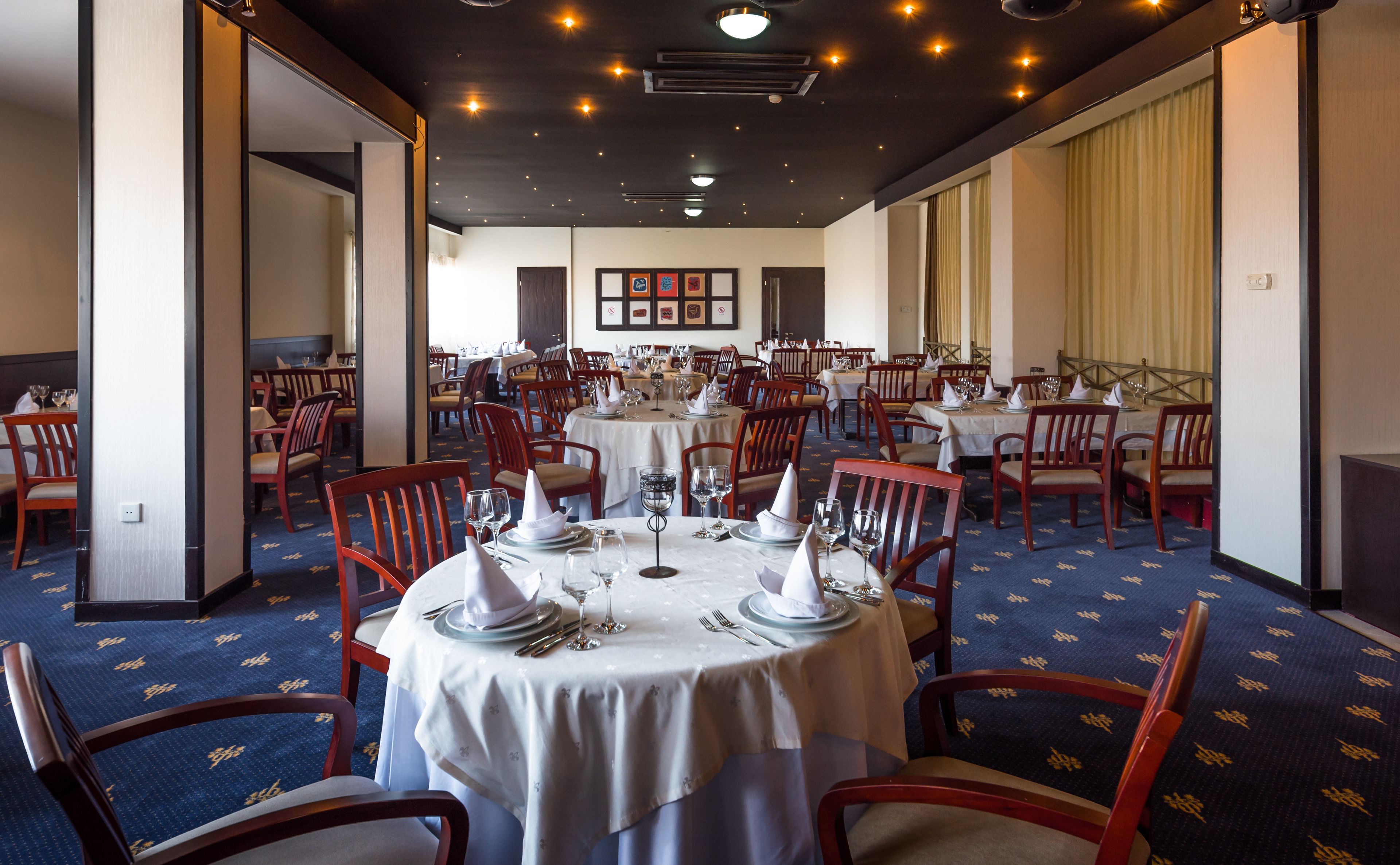 A dining area with round tables set for service, wooden chairs, and a patterned carpet.