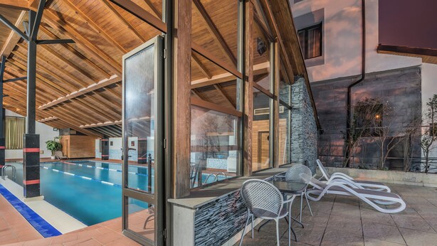 An indoor swimming pool with a glass wall, a covered patio area with seating, and a stone wall. - Kopala Tskneti (Tbilisi)