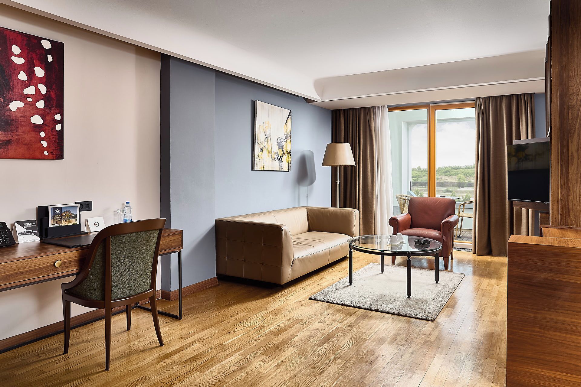 A hotel room with a sofa, armchair, coffee table, desk with chair, artwork, and a large window with curtains.