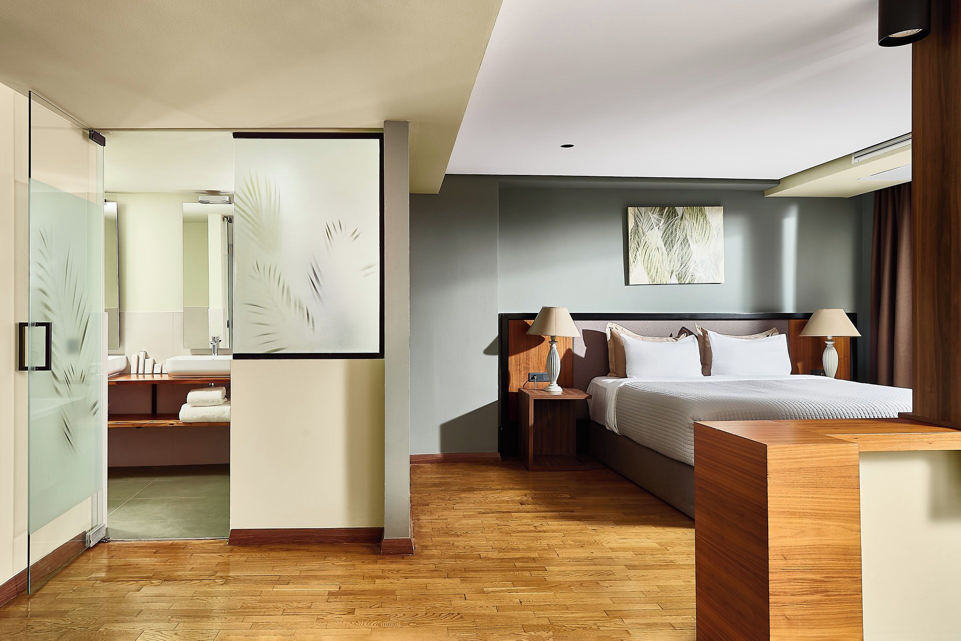 A modern hotel room with a wooden floor, a large bed with white bedding, two bedside tables with lamps, a large mirror, and a glass partition with a palm leaf design.