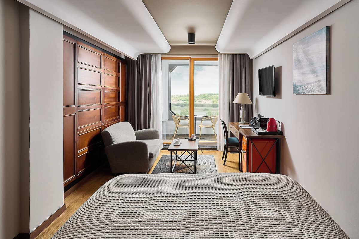 Ein Hotelzimmer mit einem großen Bett, einem Schreibtisch mit Lampe, einem Sessel und Blick ins Freie.